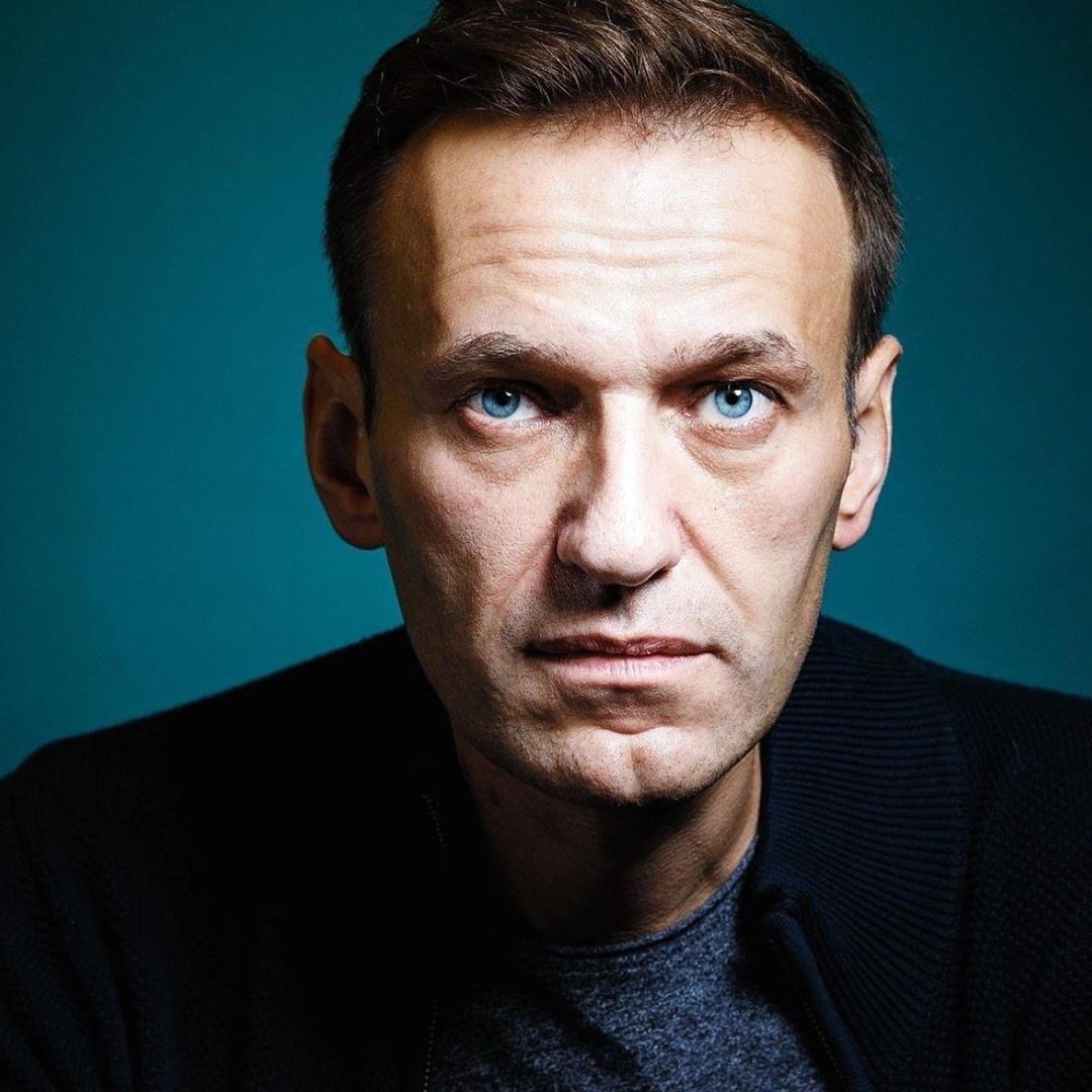 Portrait of a fair-skinned adult male with blue eyes, wearing a dark jacket over a gray shirt against a teal background. Close-up shot.