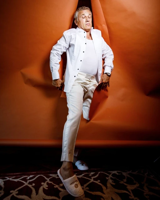 Adult man in a white shirt and pants posing while stepping through a torn orange paper, wearing slippers, with carpet visible.