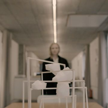 Abstract architectural model with white geometric shapes on a wooden table in a bright hallway, featuring a blurry human figure.