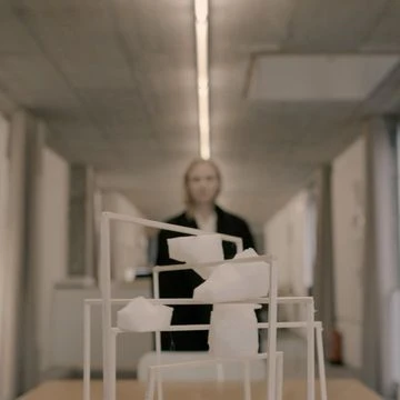 Abstract architectural model with white geometric shapes on a wooden table in a bright hallway, featuring a blurry human figure.