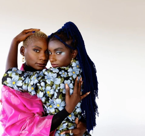 Two black women embracing closely, one with short blonde hair and one with long blue braided hair wearing a floral dress