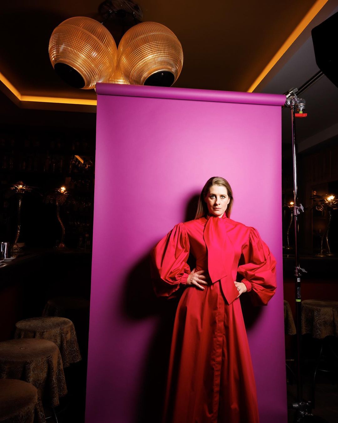 Portrait of a woman with hands on hips in a red dress, posing in front of a purple backdrop in a studio setting with modern lighting.