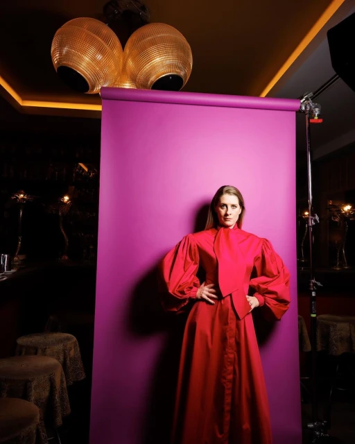 Portrait of a woman with hands on hips in a red dress, posing in front of a purple backdrop in a studio setting with modern lighting.