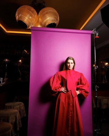 Portrait of a woman with hands on hips in a red dress, posing in front of a purple backdrop in a studio setting with modern lighting.