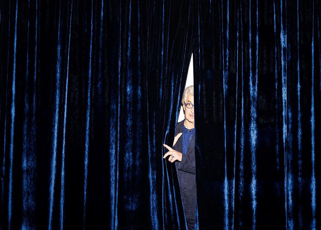 An older adult male with gray hair, wearing glasses and a navy suit, peeks through a gap in a blue velvet curtain.