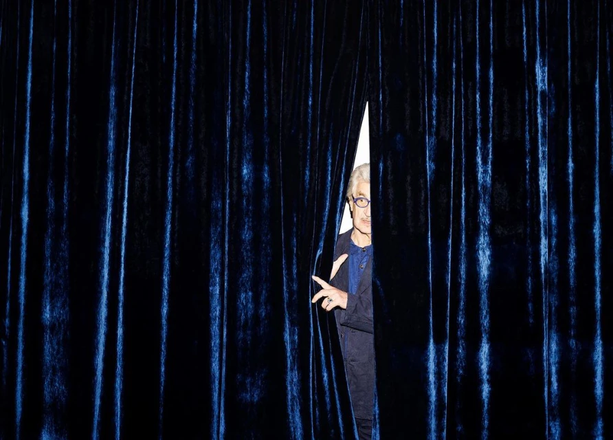 An older adult male with gray hair, wearing glasses and a navy suit, peeks through a gap in a blue velvet curtain.