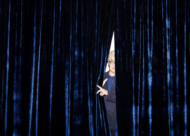 An older adult male with gray hair, wearing glasses and a navy suit, peeks through a gap in a blue velvet curtain.