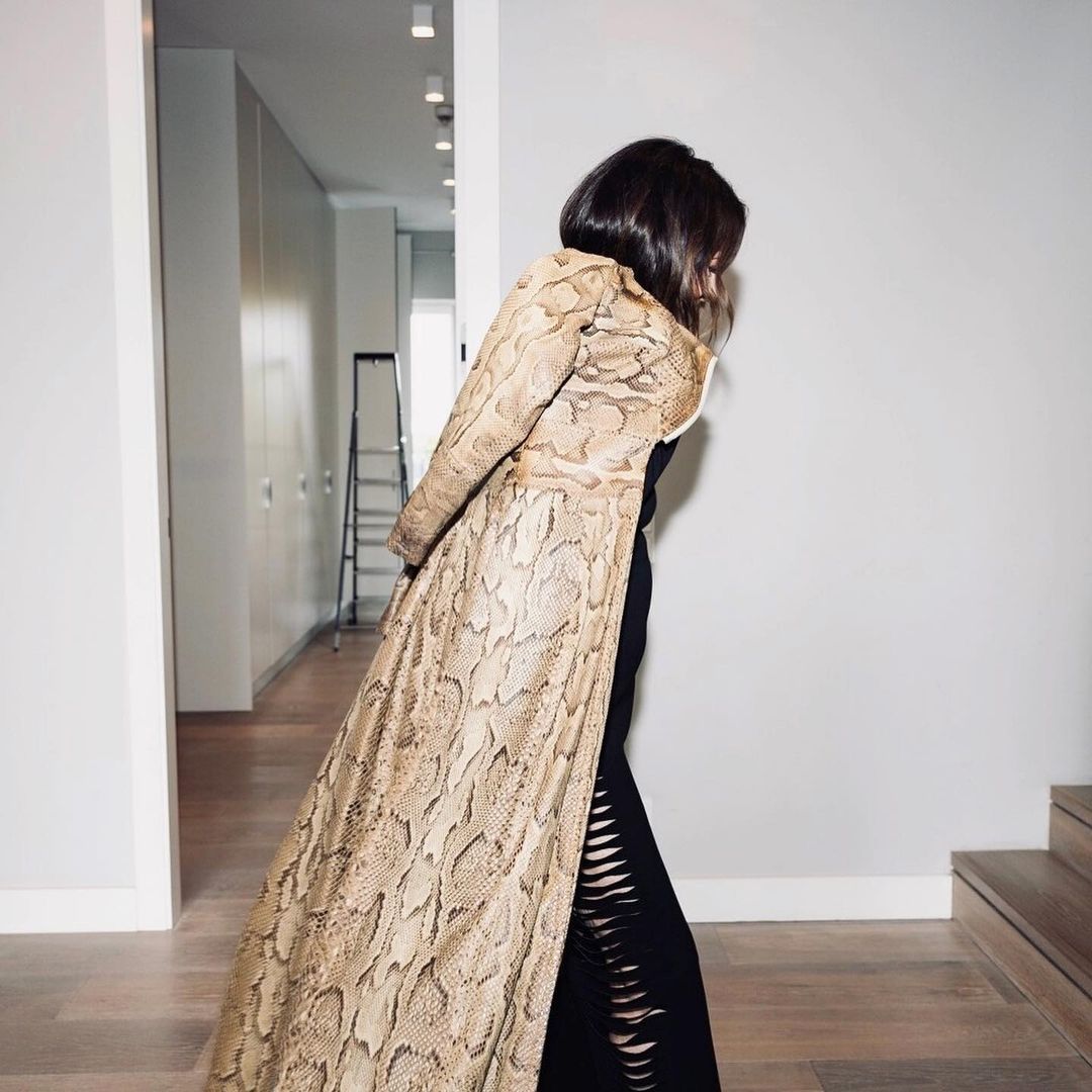 A woman in a snake-print coat and black dress poses in a hallway with white walls and a ladder in the background, evoking a fashion editorial.