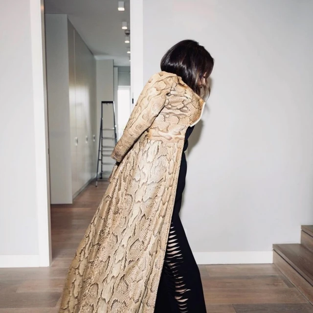 A woman in a snake-print coat and black dress poses in a hallway with white walls and a ladder in the background, evoking a fashion editorial.