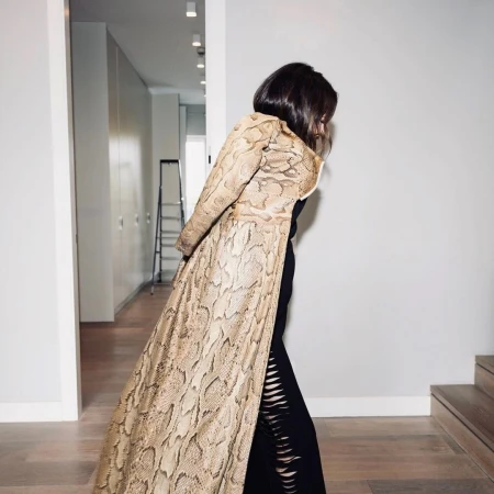 A woman in a snake-print coat and black dress poses in a hallway with white walls and a ladder in the background, evoking a fashion editorial.