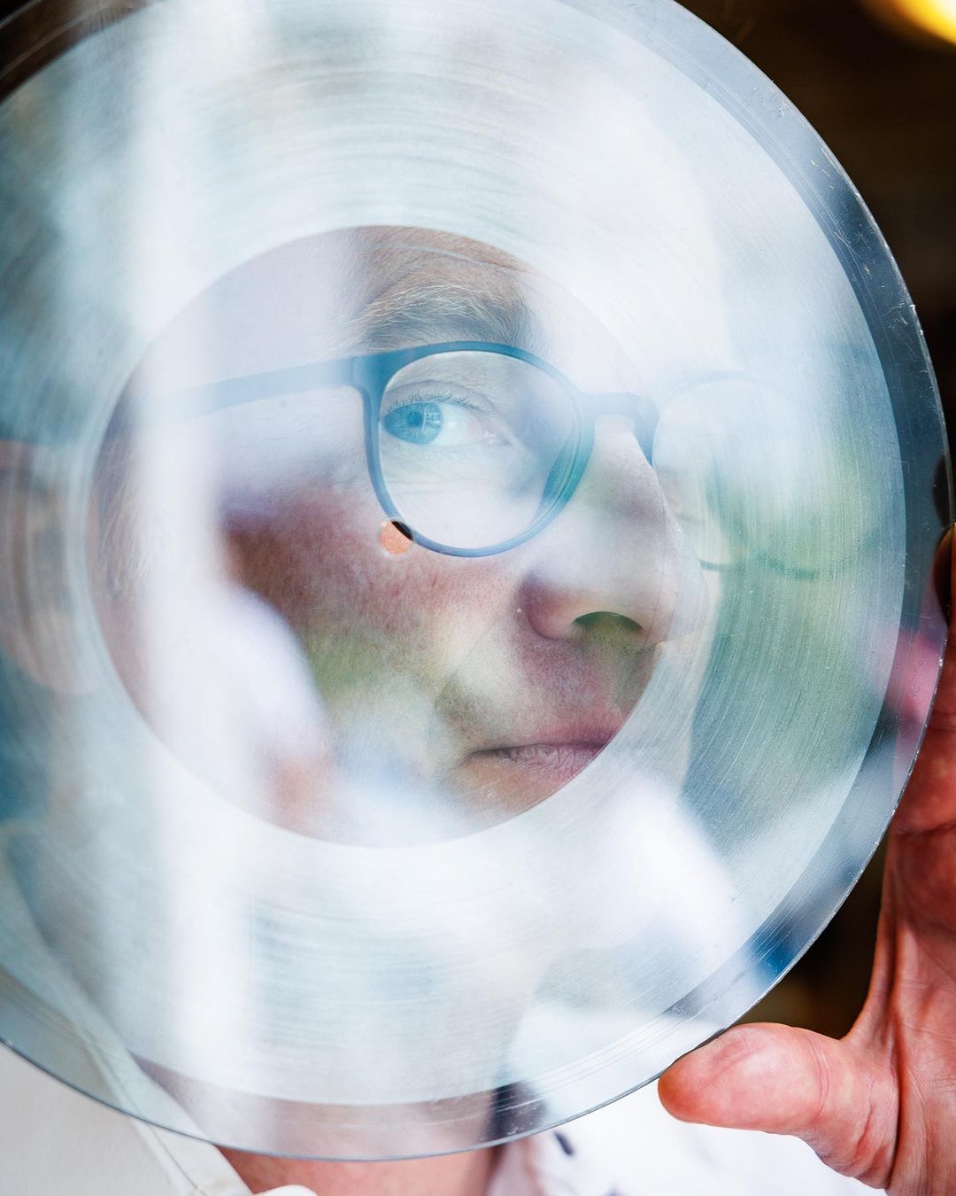 Adult male holding translucent disk with face visible through center, wearing eyeglasses in a brightly lit indoor environment.