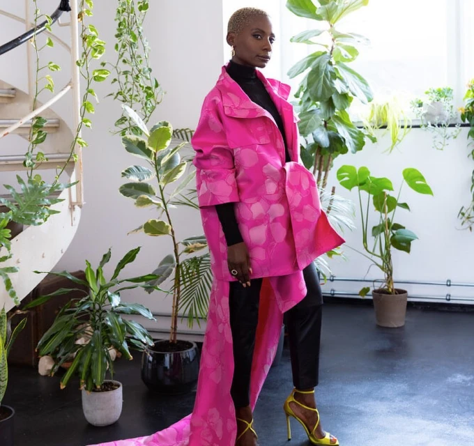 A stylish adult female with short blonde hair posing indoors wearing a bright pink floral coat, black pants, and yellow heels near plants.