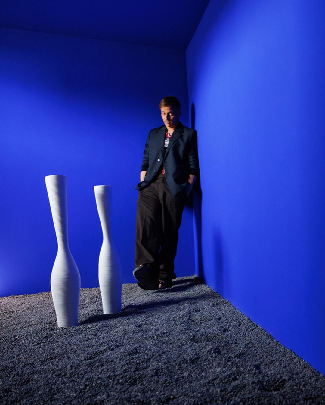 Adult male wearing blazer, leaning on bright blue wall next to two white vases on gravel floor. Fashion studio portrait with dramatic lighting.