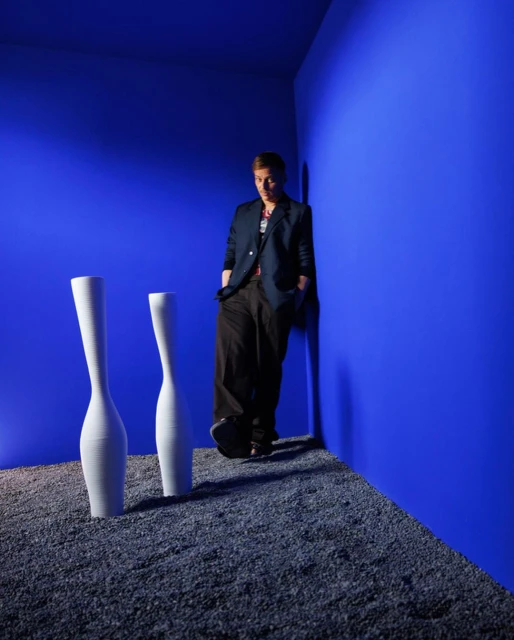 Adult male wearing blazer, leaning on bright blue wall next to two white vases on gravel floor. Fashion studio portrait with dramatic lighting.