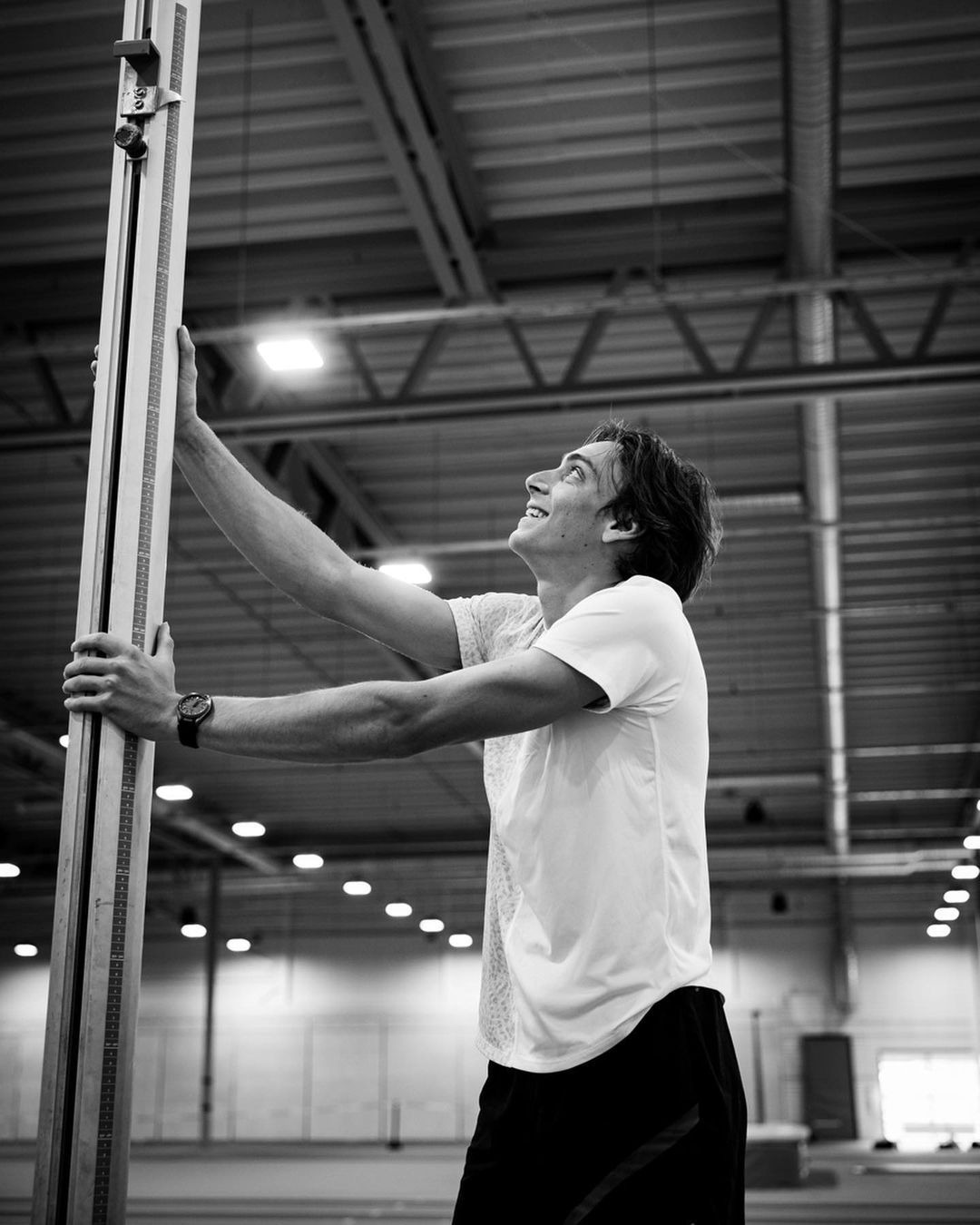 A young adult male in athletic wear adjusts a measuring pole inside a gymnasium with overhead lighting, captured in black and white.