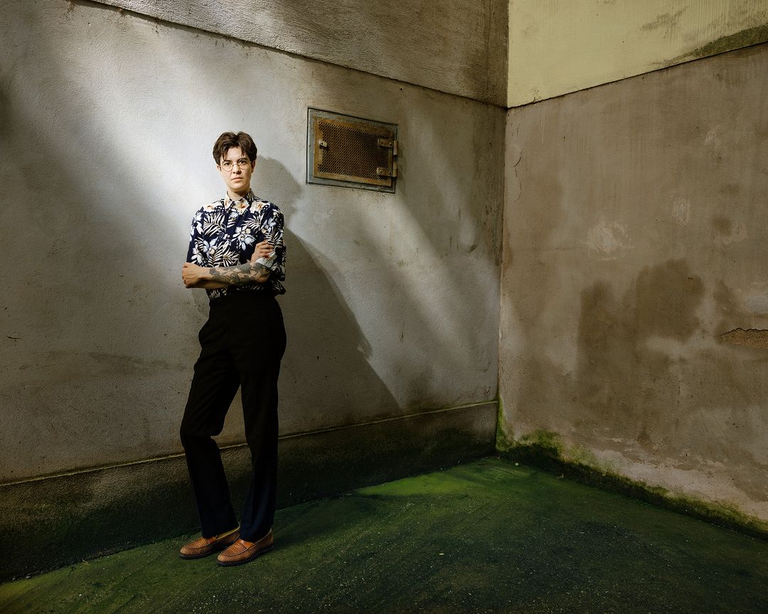 A person with crossed arms wearing a patterned shirt, dark pants, and brown shoes stands in an empty corner room with mottled walls.