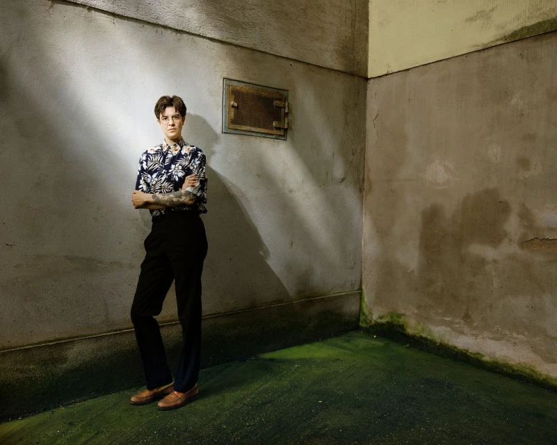 A person with crossed arms wearing a patterned shirt, dark pants, and brown shoes stands in an empty corner room with mottled walls.