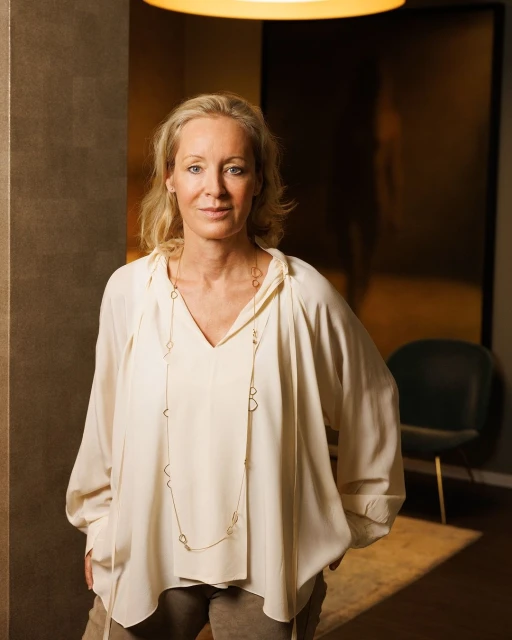 Portrait of a blonde adult woman in an off-white blouse and brown pants, posing indoors with decorative wall and soft, diffused lighting.