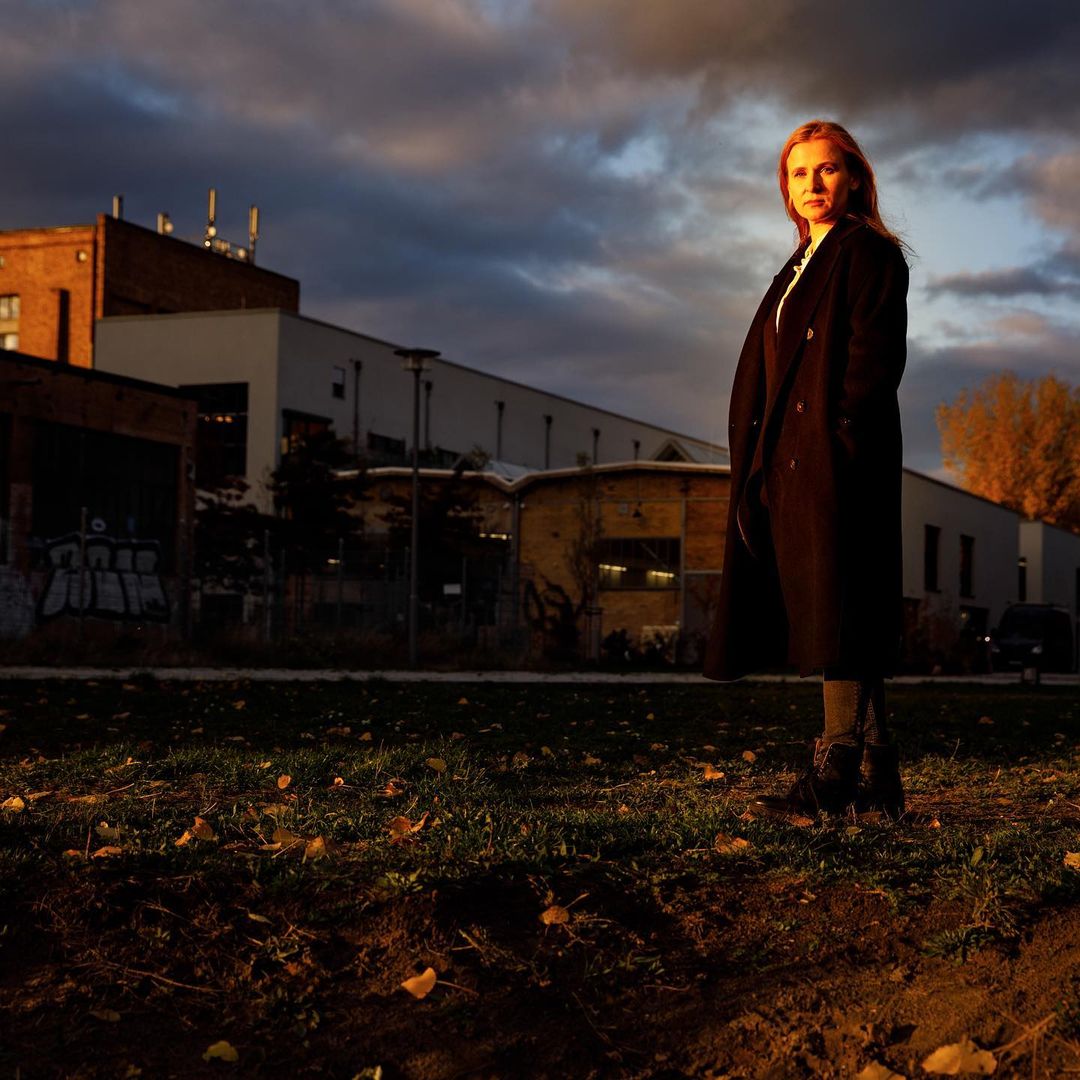 Adult woman in a long black coat stands in an urban environment with buildings and a cloudy sky at dusk. Fashion photography.