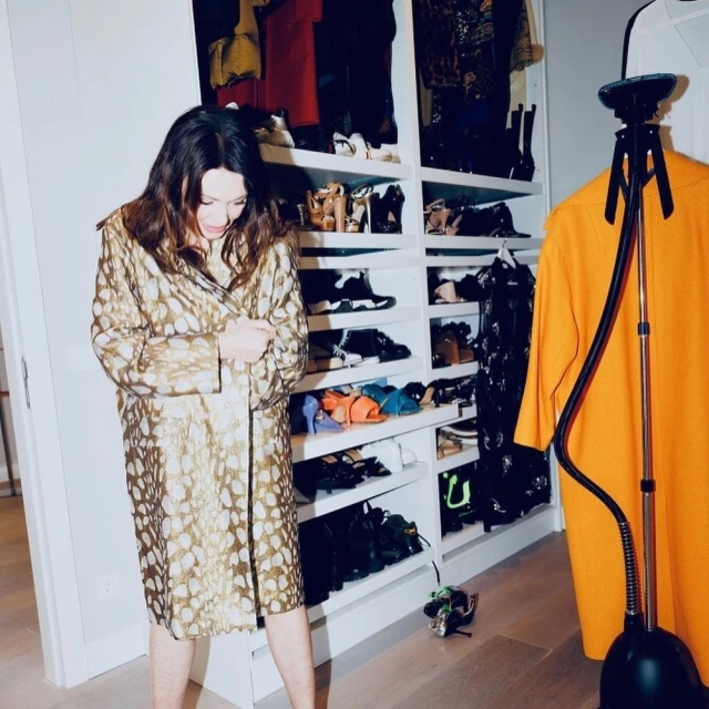 Adult woman wearing gold and tan patterned coat standing in front of a large white shelving unit with many shoes and a steamer