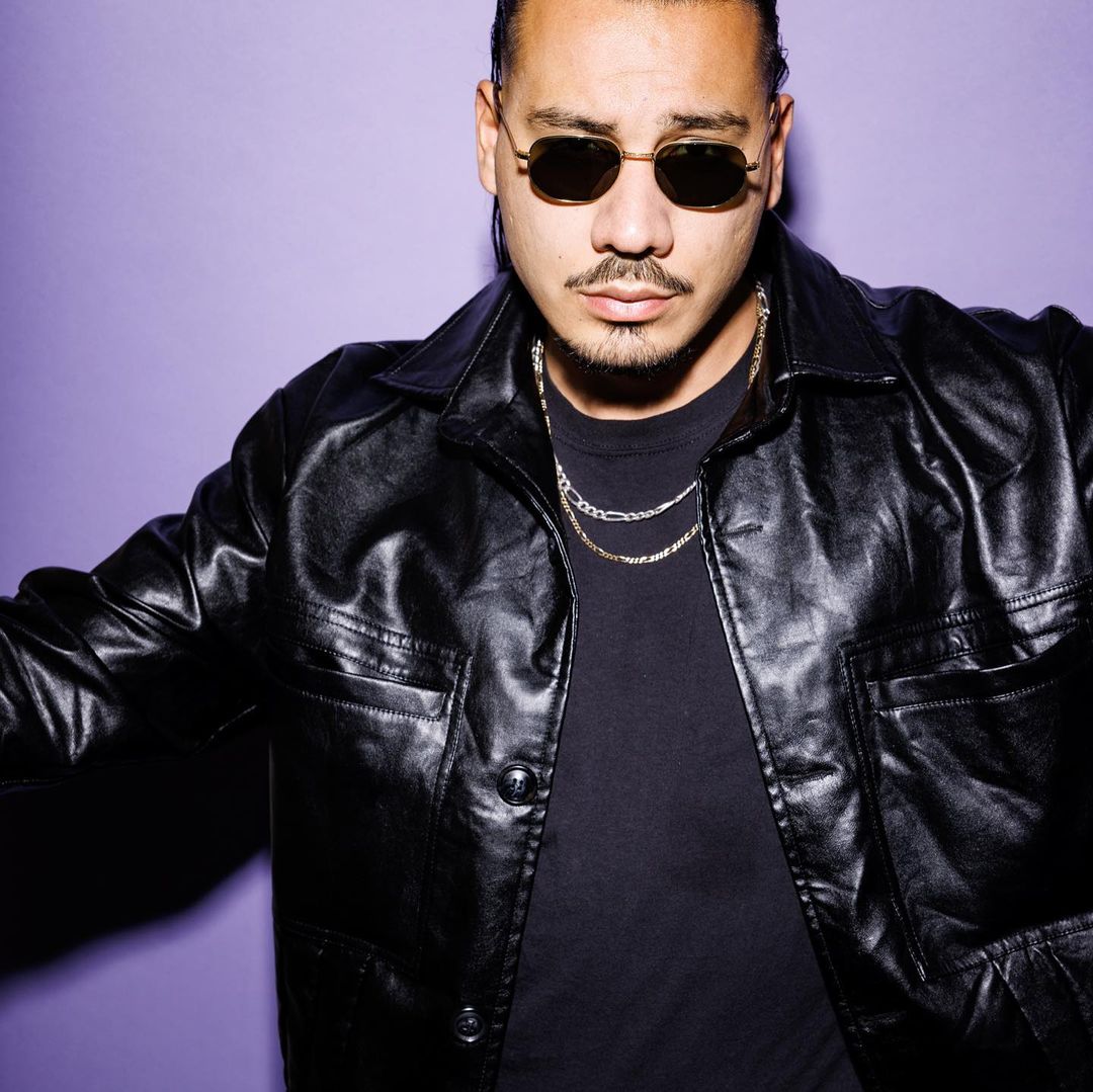 Portrait of an adult male with a goatee wearing sunglasses and a black leather jacket against a purple background in a photo studio.