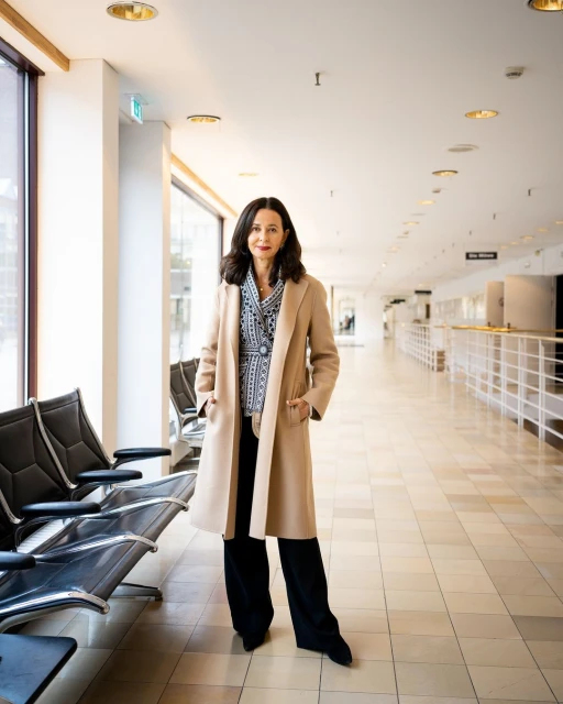 A brunette woman in a beige coat stands in a bright hallway with black benches and tile floors, looking confidently at the camera.