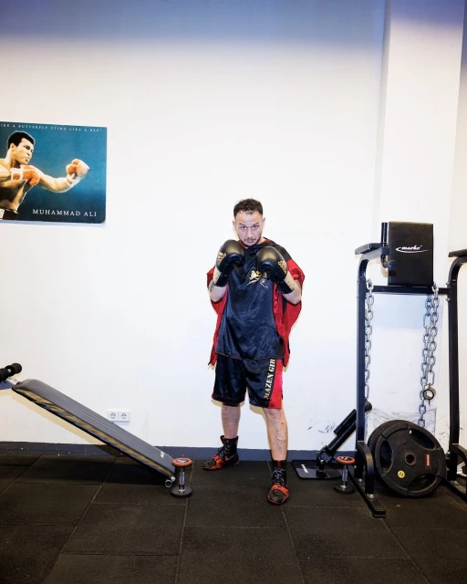 Man in boxing gear striking a pose in a gym with weights and a poster of Muhammad Ali in the background, captured in eye-level shot.