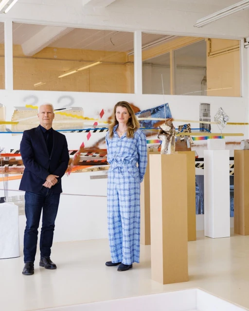 Two people pose indoors next to modern art. An adult male is on the left and an adult female is on the right wearing a plaid suit.