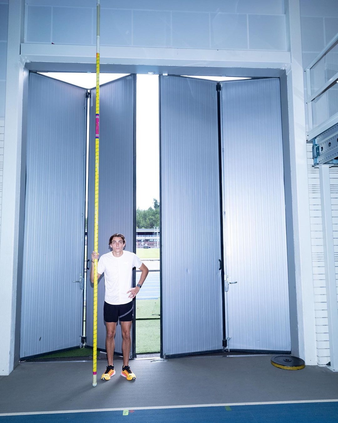 Athlete stands indoors with a yellow and pink pole vault pole in front of industrial metal doors, ready for track and field practice.