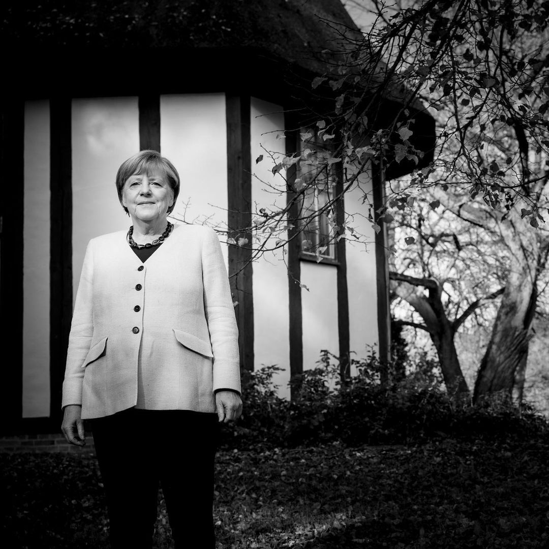 Black and white portrait of a woman in a light jacket standing outside a cottage with trees in background, captured at eye level.
