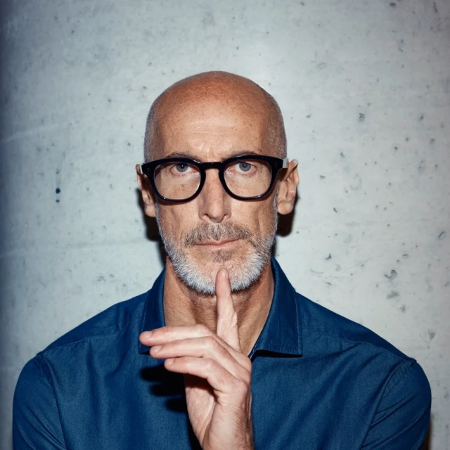 Headshot of a bald adult male with glasses and a beard, wearing a blue shirt, posing in front of a mottled gray wall with his finger to his lips.