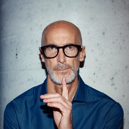 Headshot of a bald adult male with glasses and a beard, wearing a blue shirt, posing in front of a mottled gray wall with his finger to his lips.