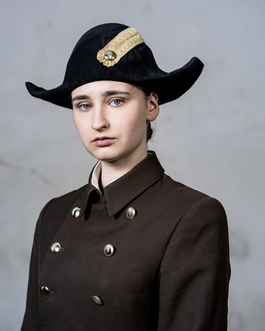 Portrait of a fair-skinned woman wearing a black tricorn hat and brown double-breasted coat in a photo studio setting.