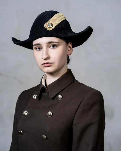 Portrait of a fair-skinned woman wearing a black tricorn hat and brown double-breasted coat in a photo studio setting.