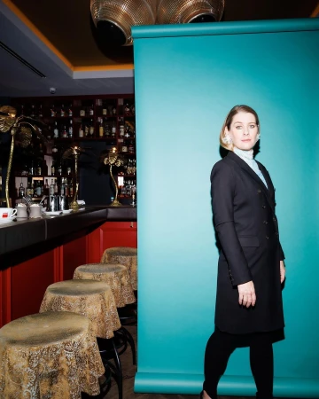 Woman in a black blazer coat standing in front of a teal backdrop inside a bar interior photographed by Peter Rigaud