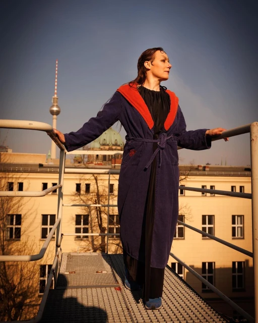 A woman in a robe standing on a rooftop with a city view, bathed in warm sunlight, showcasing an urban scene.