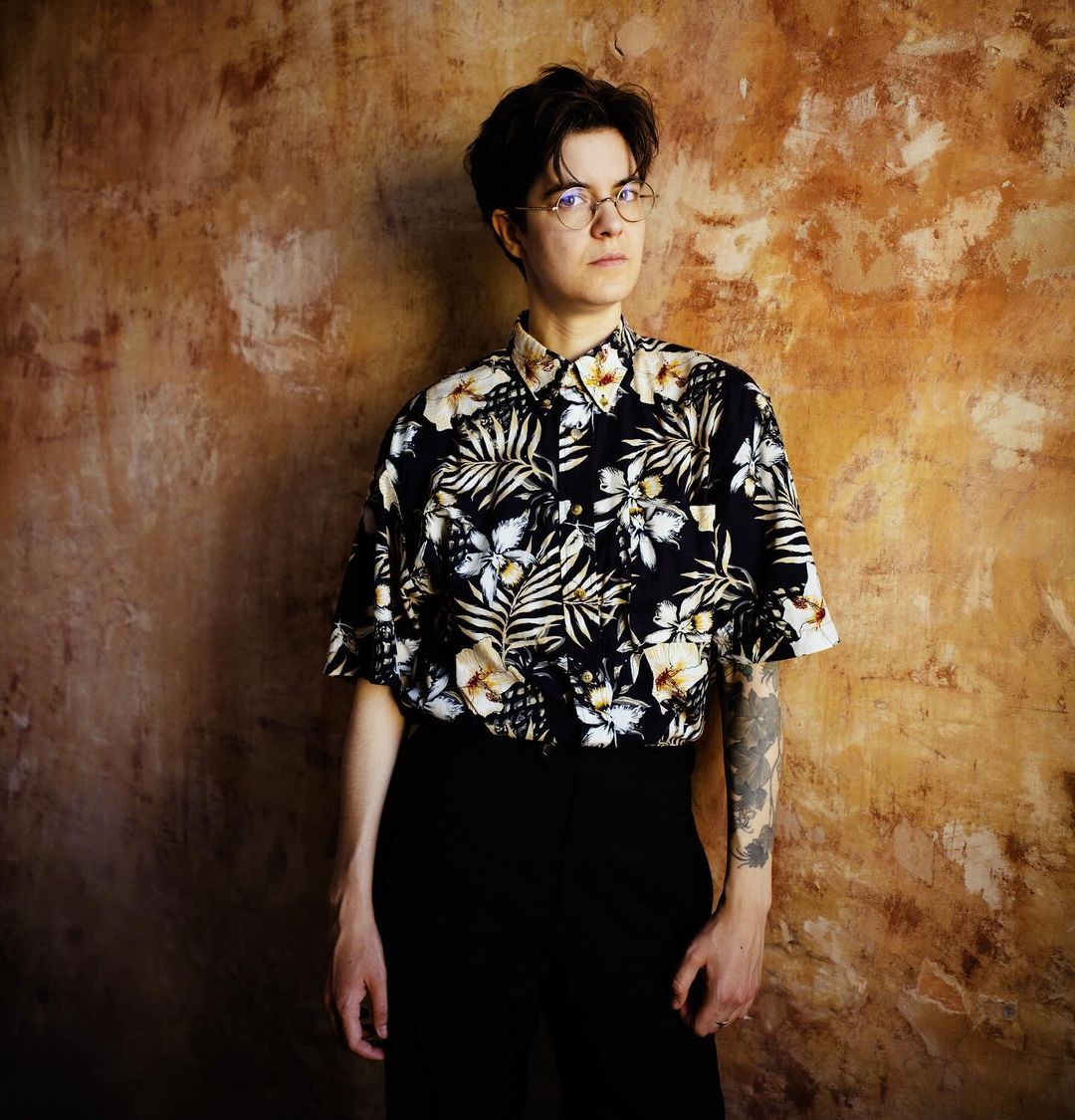 Portrait of a young adult woman with short hair and glasses wearing a floral shirt against a textured neutral-tone background.