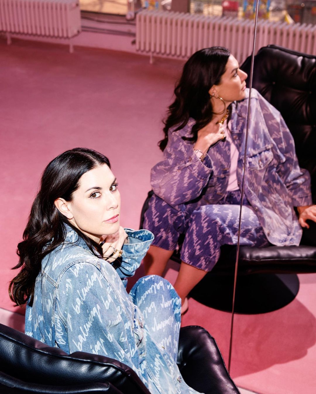 Two women pose in Balenciaga denim outfits in a pink room with black chairs and mirrors, creating a stylish and modern fashion image.