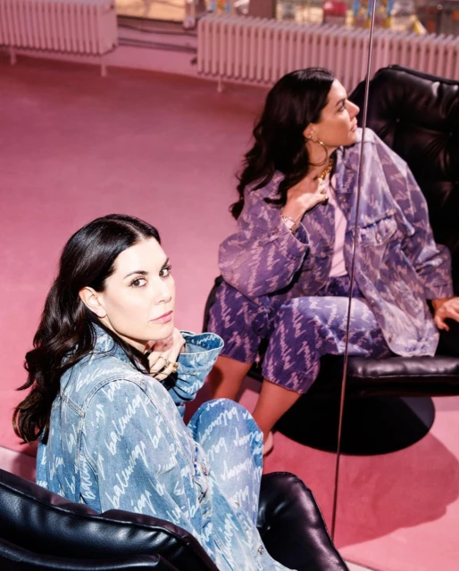 Two women pose in Balenciaga denim outfits in a pink room with black chairs and mirrors, creating a stylish and modern fashion image.