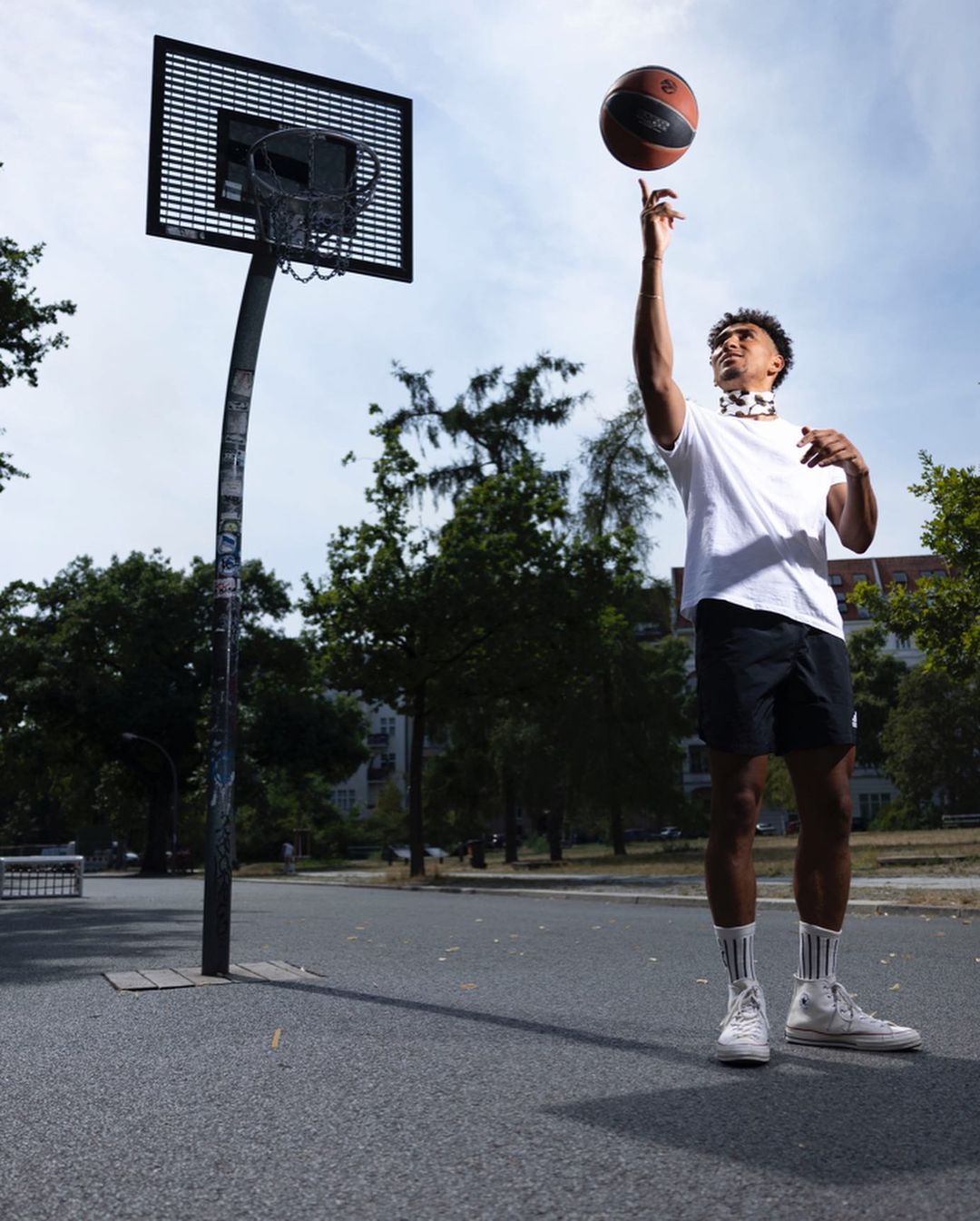 Athletic African American man in sportswear spinning a basketball on one finger, standing on an outdoor court with a basketball hoop.