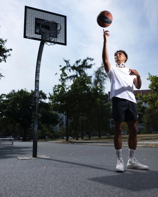 Athletic African American man in sportswear spinning a basketball on one finger, standing on an outdoor court with a basketball hoop.