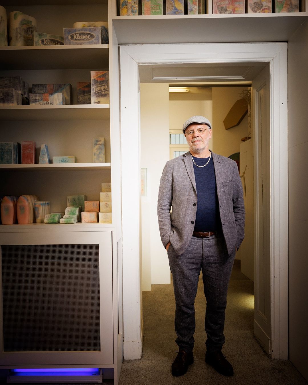 Full-length portrait of an adult man in a grey suit and cap, standing in an interior hallway with shelves of boxes and objects.