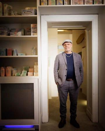 Full-length portrait of an adult man in a grey suit and cap, standing in an interior hallway with shelves of boxes and objects.