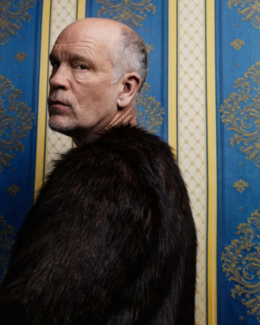 Portrait of adult male wearing a fur coat against a patterned wallpaper, with blue and gold damask and yellow vertical stripes.