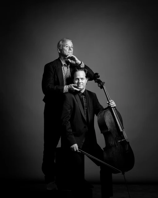 Monochrome portrait of two adult men in suits, one seated holding a cello bow, the other standing with hands near his face.