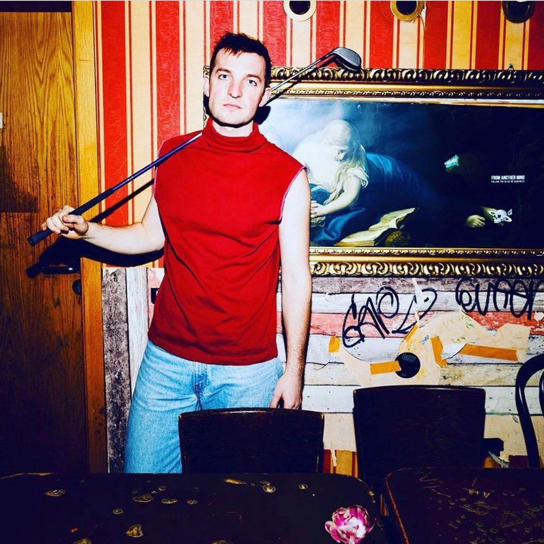 Man in a red turtleneck and jeans poses with a golf club in an eclectic interior with artwork, wallpaper, and graffiti details.