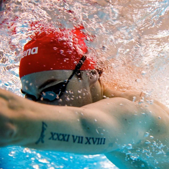Underwater close-up of a swimmer wearing a red swim cap and goggles, showing an arm tattoo, surrounded by water and bubbles.