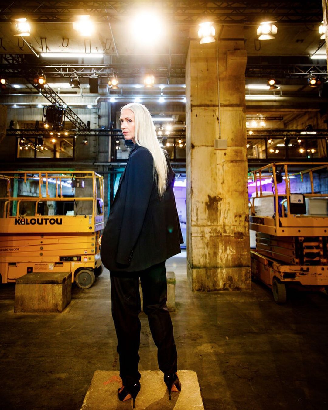 A woman with long platinum hair poses in a black blazer and pants, set against an industrial background with scissor lifts and concrete columns.