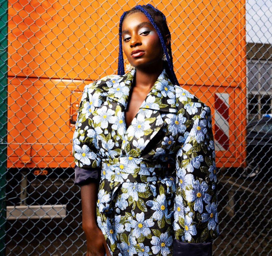 Stylish woman in a floral trench coat poses in front of an orange fence, showcasing a modern fashion aesthetic and bold color contrast.