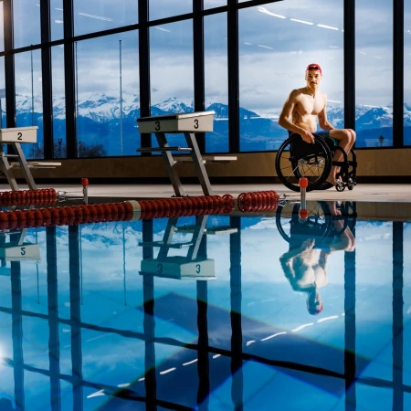 Man in wheelchair at indoor pool, mountains and natural light backdrop, reflections in water, swimming pool starting blocks visible.